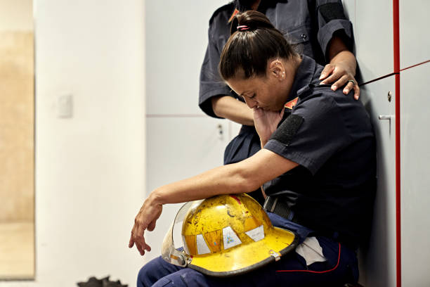 Side view of late 30s woman in uniform sitting on bench in fire station locker room with helmet in lap, head down, and eyes closed.